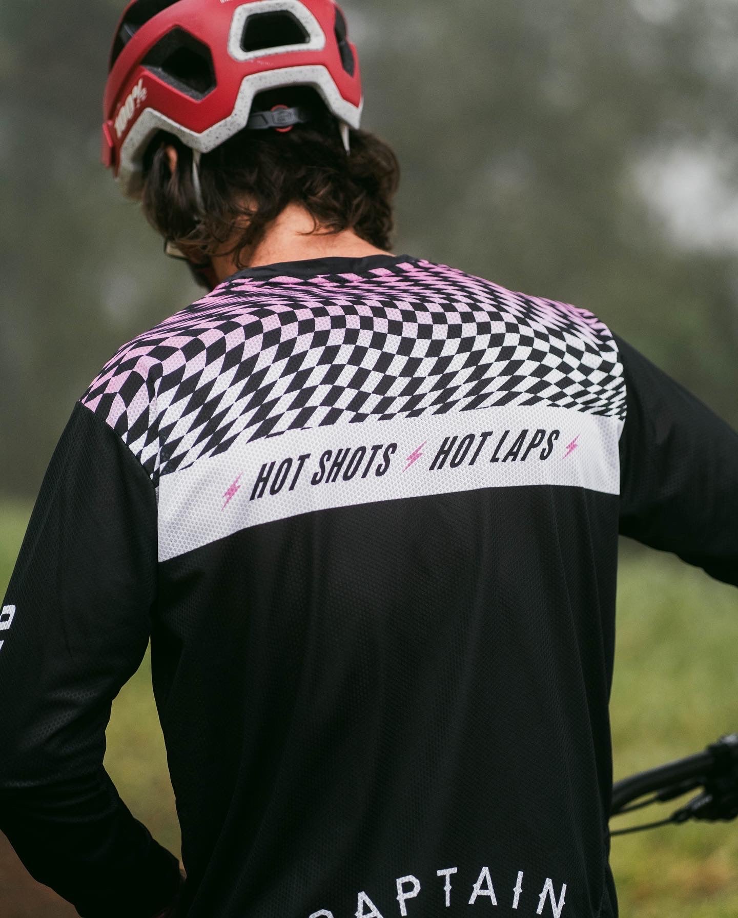 A biker wears the Captain and Stoker MTB Jersey. It has a black, white, and pink wavy checkerboard design on the front and back of the shoulders, with a white bar containing the words "Hot Shots, Hot Laps" across the back. 