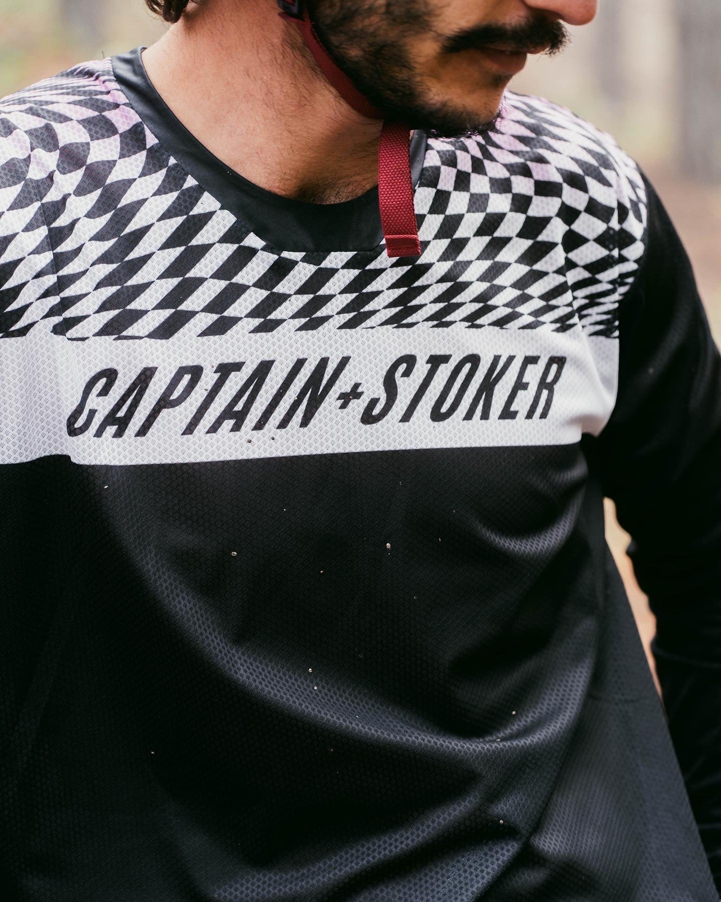 A biker wears the Captain and Stoker MTB Jersey. It has a black, white, and pink wavy checkerboard design on the front and back of the shoulders, with a white bar containing the words "Captain and Stoker" across the chest. 