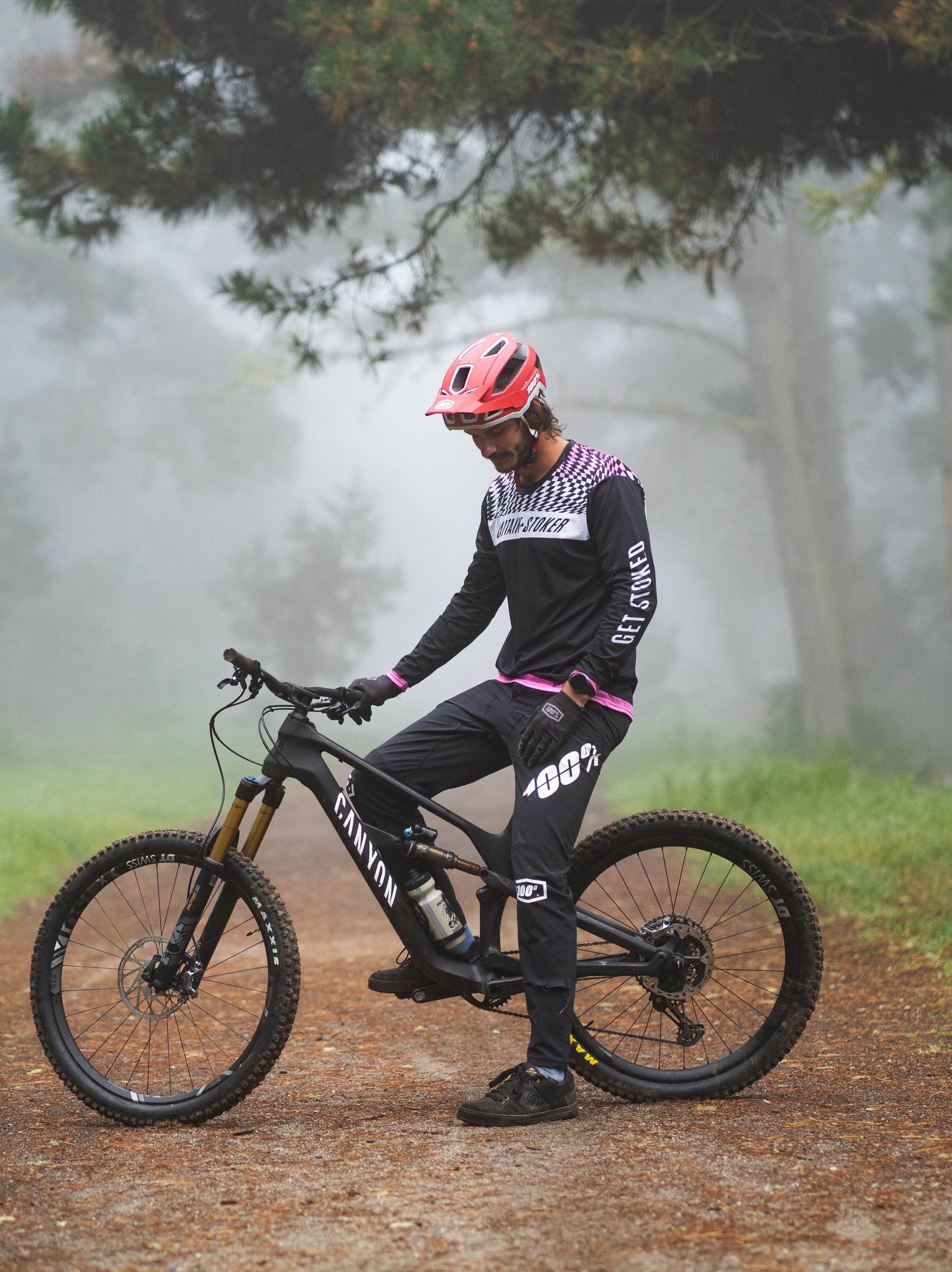 A biker wears the black Captain and Stoker MTB Jersey while riding a bike. It has a black, white, and pink wavy checkerboard design on the front and back of the shoulders. 