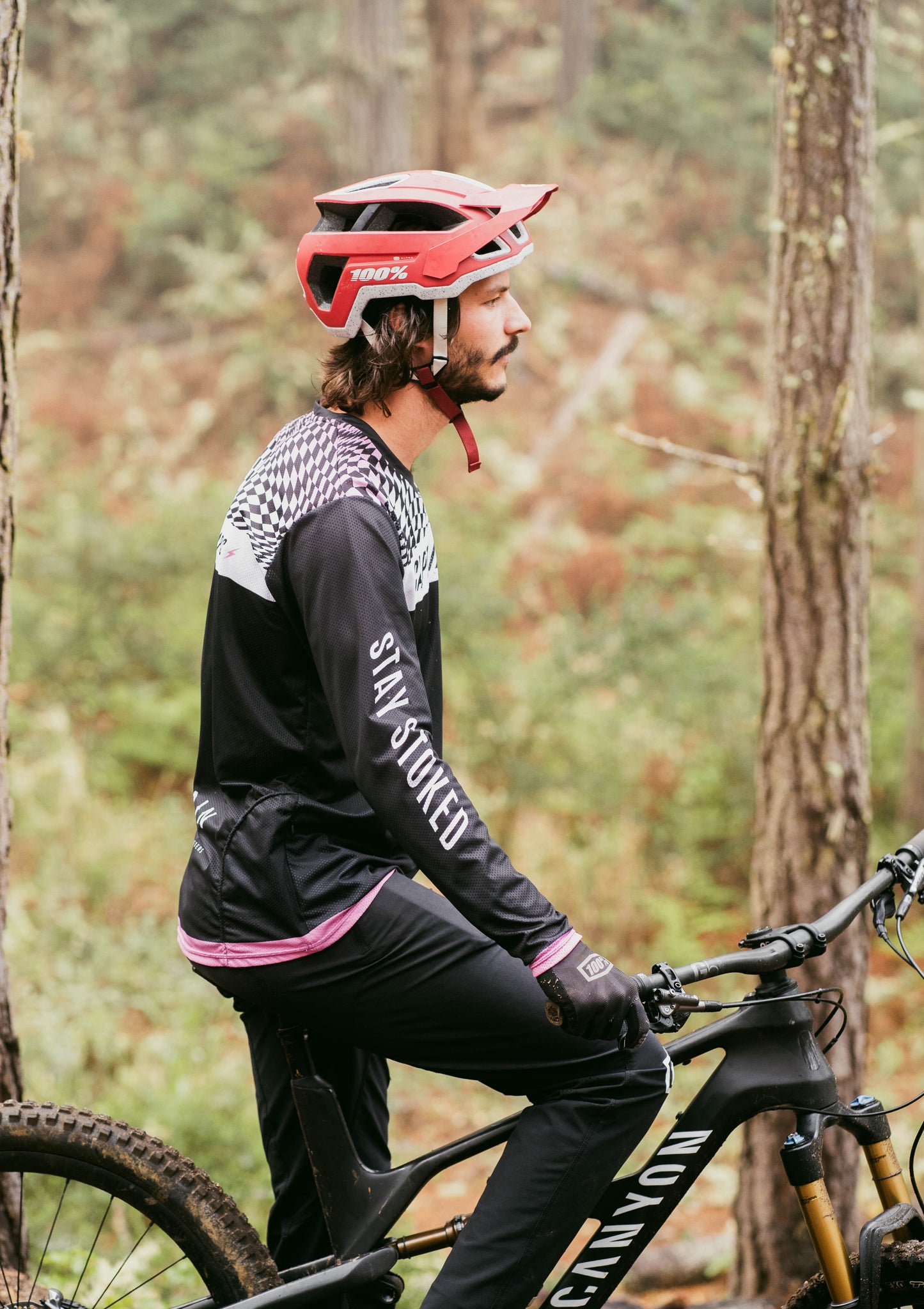 A biker wears the Captain and Stoker MTB Jersey while riding a bike. It has a black, white, and pink wavy checkerboard design on the front and back of the shoulders, with the text "Get Stoked" down the arm. 